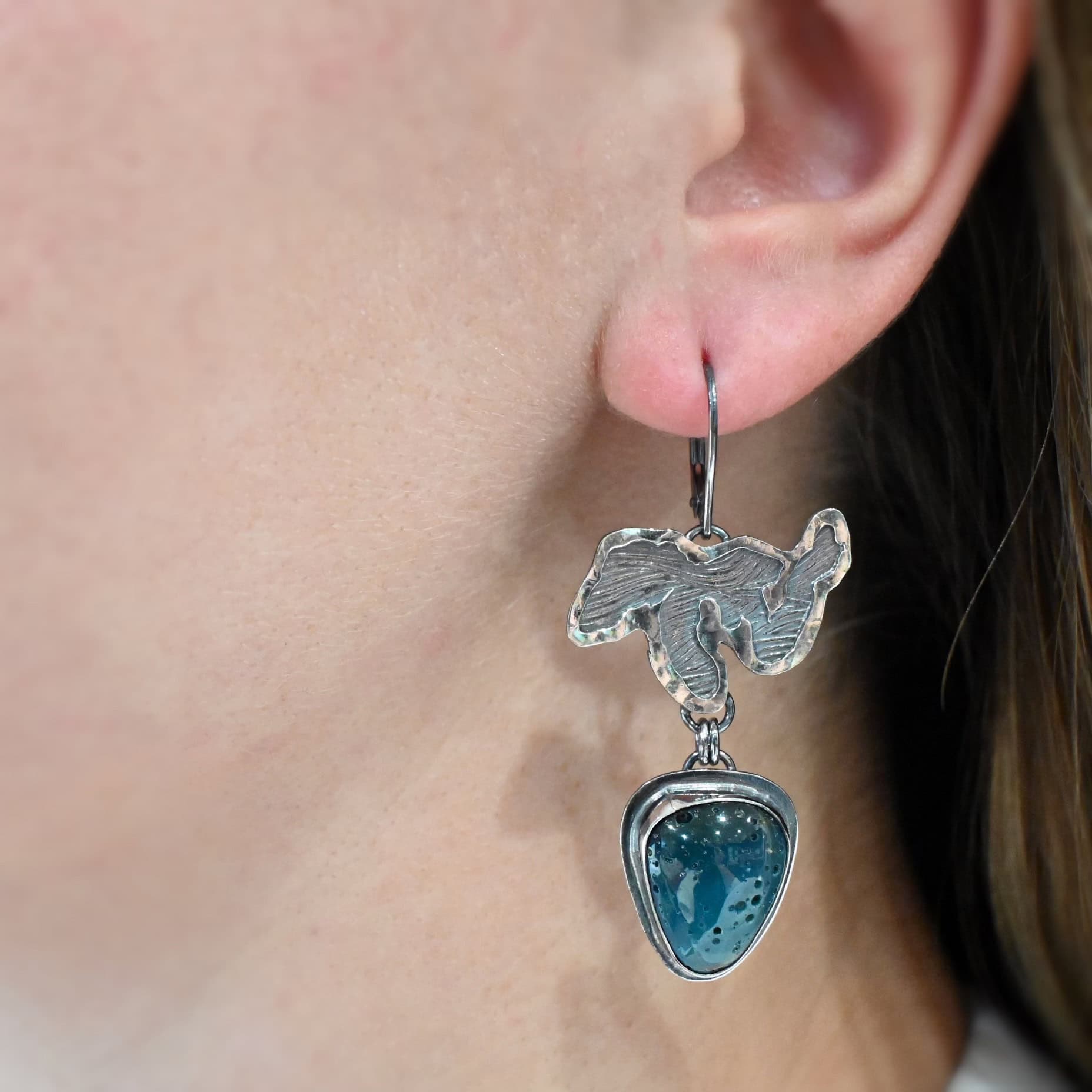 Close up of the "Lake Superior" Earring as shown on a model. Made out of sterling silver and leland blue. Earring has a sterling silver cut out that looks similar to lake superior on the top of it with a leland blue stone underneath it. The lake has a loop on the bottom back of them in which two jump rings are connected to the top of the loop on the back of the leland blue. There is a leland blue stone that is teardrop shaped. Earring is on leverback ear wires and are worn by a model to demonstrate length.