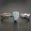 Close up of the "Water Contained" Bracelet. Made out of sterling silver and aquamarine. The aquamarine is an oval shape, soldered to the cuff. The cuff is wavy, and has a thick solid sterling silver line that is flat, but raised on both sides. There are silver dots that are scattered all over the bracelet. Bracelet is set against a dark background.