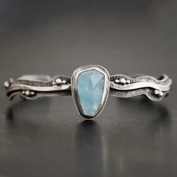 Close up of the "Water Contained" Bracelet. Made out of sterling silver and aquamarine. The aquamarine is an oval shape, soldered to the cuff. The cuff is wavy, and has a thick solid sterling silver line that is flat, but raised on both sides. There are silver dots that are scattered all over the bracelet. Bracelet is set against a dark background.