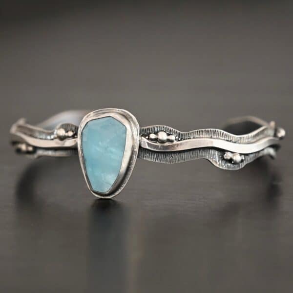 Close up of the "Water Contained" Bracelet. Made out of sterling silver and aquamarine. The aquamarine is an oval shape, soldered to the cuff. The cuff is wavy, and has a thick solid sterling silver line that is flat, but raised on both sides. There are silver dots that are scattered all over the bracelet. Bracelet is set against a dark background.