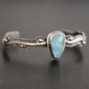 Close up of the "Water Contained" Bracelet. Made out of sterling silver and aquamarine. The aquamarine is an oval shape, soldered to the cuff. The cuff is wavy, and has a thick solid sterling silver line that is flat, but raised on both sides. There are silver dots that are scattered all over the bracelet. Bracelet is set against a dark background.