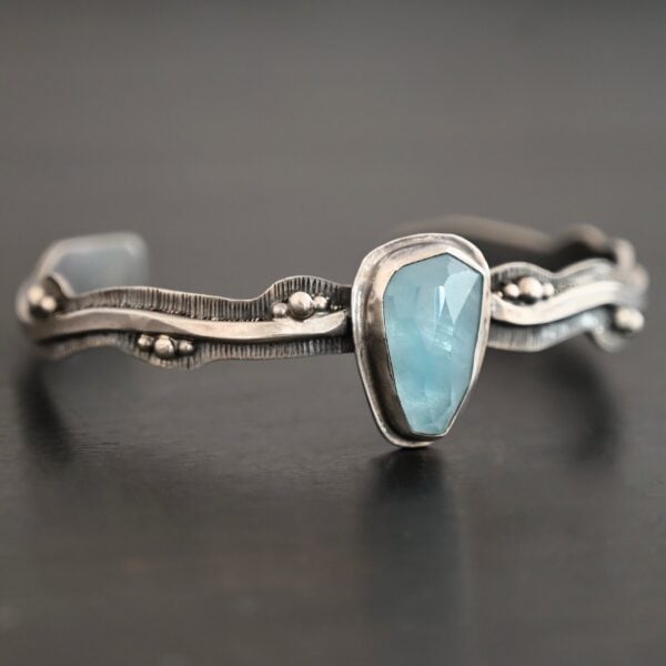 Close up of the "Water Contained" Bracelet. Made out of sterling silver and aquamarine. The aquamarine is an oval shape, soldered to the cuff. The cuff is wavy, and has a thick solid sterling silver line that is flat, but raised on both sides. There are silver dots that are scattered all over the bracelet. Bracelet is set against a dark background.