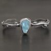 Close up of the "Water Contained" Bracelet. Made out of sterling silver and aquamarine. The aquamarine is an oval shape, soldered to the cuff. The cuff is wavy, and has a thick solid sterling silver line that is flat, but raised on both sides. There are silver dots that are scattered all over the bracelet. Bracelet is set against a dark background.