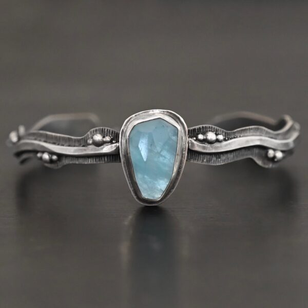 Close up of the "Water Contained" Bracelet. Made out of sterling silver and aquamarine. The aquamarine is an oval shape, soldered to the cuff. The cuff is wavy, and has a thick solid sterling silver line that is flat, but raised on both sides. There are silver dots that are scattered all over the bracelet. Bracelet is set against a dark background.
