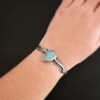 Close up of the "Water Contained" Bracelet. Made out of sterling silver and aquamarine. The aquamarine is an oval shape, soldered to the cuff. The cuff is wavy, and has a thick solid sterling silver line that is flat, but raised on both sides. There are silver dots that are scattered all over the bracelet. Bracelet is worn by a model on their wrist to demonstrate the size of the stone and the thickness of the cuff bracelet.