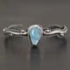 Close up of the "Water Contained" Bracelet. Made out of sterling silver and aquamarine. The aquamarine is an oval shape, soldered to the cuff. The cuff is wavy, and has a thick solid sterling silver line that is flat, but raised on both sides. There are silver dots that are scattered all over the bracelet. Bracelet is set against a dark background.
