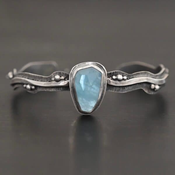 Close up of the "Water Contained" Bracelet. Made out of sterling silver and aquamarine. The aquamarine is an oval shape, soldered to the cuff. The cuff is wavy, and has a thick solid sterling silver line that is flat, but raised on both sides. There are silver dots that are scattered all over the bracelet. Bracelet is set against a dark background.