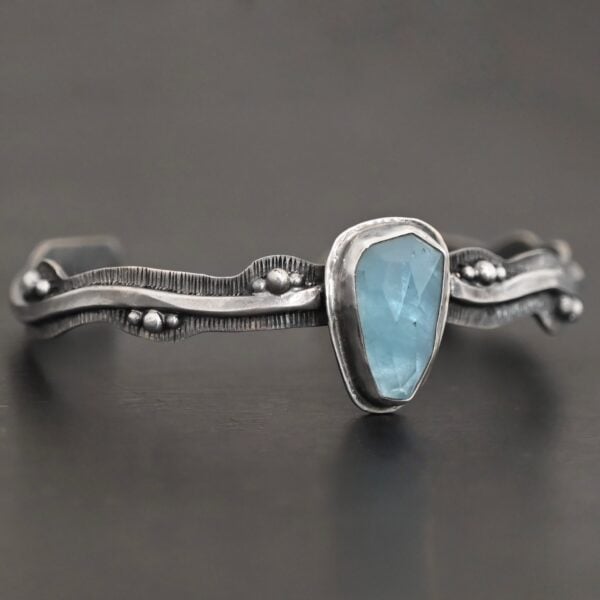 Close up of the "Water Contained" Bracelet. Made out of sterling silver and aquamarine. The aquamarine is an oval shape, soldered to the cuff. The cuff is wavy, and has a thick solid sterling silver line that is flat, but raised on both sides. There are silver dots that are scattered all over the bracelet. Bracelet is set against a dark background.