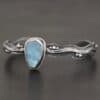 Close up of the "Water Contained" Bracelet. Made out of sterling silver and aquamarine. The aquamarine is an oval shape, soldered to the cuff. The cuff is wavy, and has a thick solid sterling silver line that is flat, but raised on both sides. There are silver dots that are scattered all over the bracelet. Bracelet is set against a dark background.