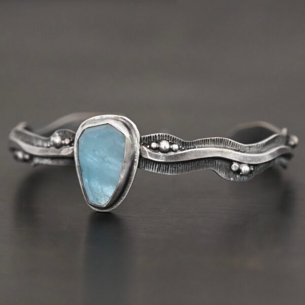 Close up of the "Water Contained" Bracelet. Made out of sterling silver and aquamarine. The aquamarine is an oval shape, soldered to the cuff. The cuff is wavy, and has a thick solid sterling silver line that is flat, but raised on both sides. There are silver dots that are scattered all over the bracelet. Bracelet is set against a dark background.
