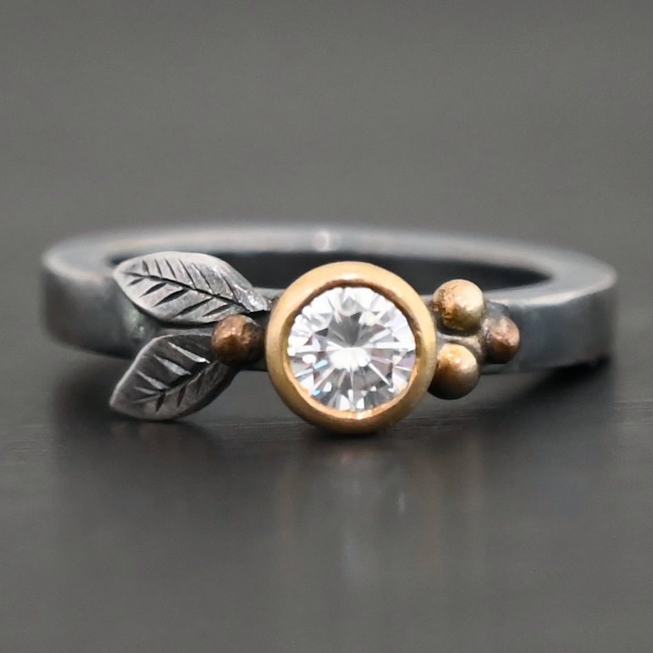 Here's To Forever Ring New "Here's To Forever" Ring as shown up close. Ring is made out of sterling silver, gold, and moissanite. There are two sterling silver engraved leaves on the left side of the ring with a gold ball at the end of them, and closest to the moissanite. There are three gold balls on the right side of the ring. In the center of the ring is a moissanite that is set in gold. Ring is set against a dark background.