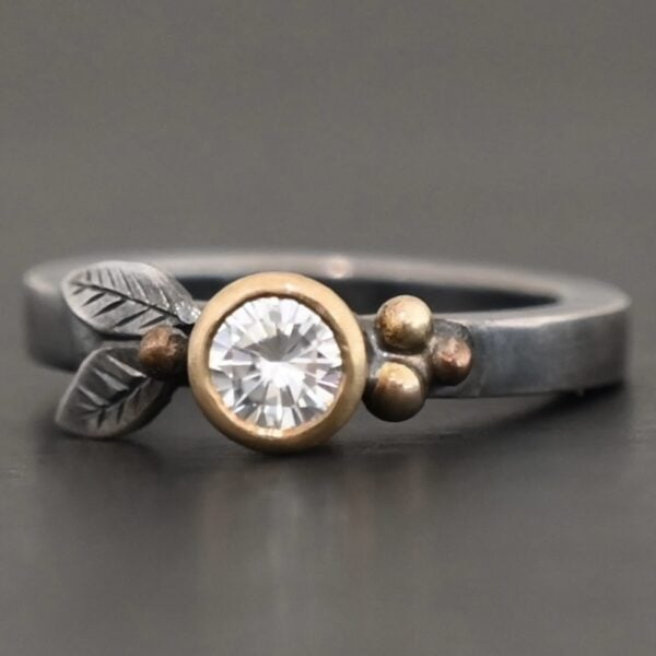 "Here's To Forever" Ring as shown up close. Ring is made out of sterling silver, gold, and moissanite. There are two sterling silver engraved leaves on the left side of the ring with a gold ball at the end of them, and closest to the moissanite. There are three gold balls on the right side of the ring. In the center of the ring is a moissanite that is set in gold. Ring is set against a dark background.
