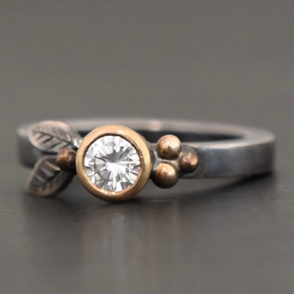 "Here's To Forever" Ring as shown up close. Ring is made out of sterling silver, gold, and moissanite. There are two sterling silver engraved leaves on the left side of the ring with a gold ball at the end of them, and closest to the moissanite. There are three gold balls on the right side of the ring. In the center of the ring is a moissanite that is set in gold. Ring is set against a dark background.