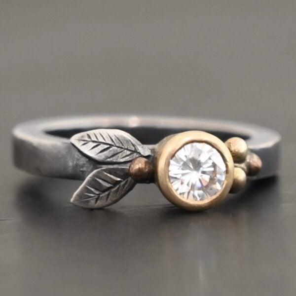 "Here's To Forever" Ring as shown up close. Ring is made out of sterling silver, gold, and moissanite. There are two sterling silver engraved leaves on the left side of the ring with a gold ball at the end of them, and closest to the moissanite. There are three gold balls on the right side of the ring. In the center of the ring is a moissanite that is set in gold. Ring is set against a dark background.