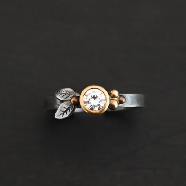 "Here's To Forever" Ring as shown up close. Ring is made out of sterling silver, gold, and moissanite. There are two sterling silver engraved leaves on the left side of the ring with a gold ball at the end of them, and closest to the moissanite. There are three gold balls on the right side of the ring. In the center of the ring is a moissanite that is set in gold. Ring is set against a dark background.