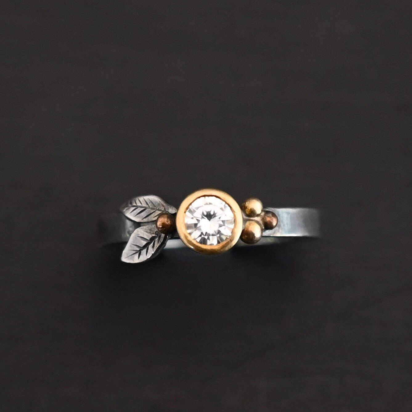 Here's To Forever Ring New "Here's To Forever" Ring as shown up close. Ring is made out of sterling silver, gold, and moissanite. There are two sterling silver engraved leaves on the left side of the ring with a gold ball at the end of them, and closest to the moissanite. There are three gold balls on the right side of the ring. In the center of the ring is a moissanite that is set in gold. Ring is set against a dark background.