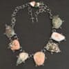 The "Look For Adventure" Necklace. Made out of sterling silver and ocean jasper. There are nine ocean jasper stones that create this necklace. Seven of them are large stones, and there are two smaller ones on the back that are a part of the clasp for the necklace. Each stone is a various shape, size, and color from one another. There is a lot of green and orange colors, with a bit of red in some of the stones. Each ocean jasper has multiple sterling silver dots going around the stone setting. The chain is made out of sterling silver hammered circular links that are oxidized. Necklace is sitting against a dark background.