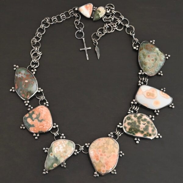 The "Look For Adventure" Necklace. Made out of sterling silver and ocean jasper. There are nine ocean jasper stones that create this necklace. Seven of them are large stones, and there are two smaller ones on the back that are a part of the clasp for the necklace. Each stone is a various shape, size, and color from one another. There is a lot of green and orange colors, with a bit of red in some of the stones. Each ocean jasper has multiple sterling silver dots going around the stone setting. The chain is made out of sterling silver hammered circular links that are oxidized. Necklace is sitting against a dark background.