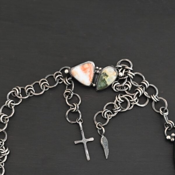The "Look For Adventure" Necklace clasp. Made out of sterling silver and ocean jasper. There are two ocean jasper that make up the clasp. One is teardrop shaped and the other is triangle shaped. The teardrop shaped ocean jasper is on the left side of the clasp. It is dark green from the tip of the teardrop to the middle of the stone. The bottom half of the stone is yellow with white spots. The triangle shaped ocean jasper is on the right side of the clasp. It is orange from the top point of the triangle to the center. The bottom half of the triangle is white. The clasp is attached to a handmade sterling silver chain that has large circular loops that are hammered and oxidized. The circle loops are attached together with jump rings. Clasp is sitting against a dark background.