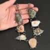The "Look For Adventure" Necklace. Made out of sterling silver and ocean jasper. There are nine ocean jasper stones that create this necklace. Seven of them are large stones, and there are two smaller ones on the back that are a part of the clasp for the necklace. Each stone is a various shape, size, and color from one another. There is a lot of green and orange colors, with a bit of red in some of the stones. Each ocean jasper has multiple sterling silver dots going around the stone setting. The chain is made out of sterling silver hammered circular links that are oxidized. Necklace is sitting against a dark background.