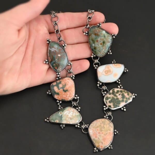 The "Look For Adventure" Necklace. Made out of sterling silver and ocean jasper. There are nine ocean jasper stones that create this necklace. Seven of them are large stones, and there are two smaller ones on the back that are a part of the clasp for the necklace. Each stone is a various shape, size, and color from one another. There is a lot of green and orange colors, with a bit of red in some of the stones. Each ocean jasper has multiple sterling silver dots going around the stone setting. The chain is made out of sterling silver hammered circular links that are oxidized. Necklace is sitting against a dark background.