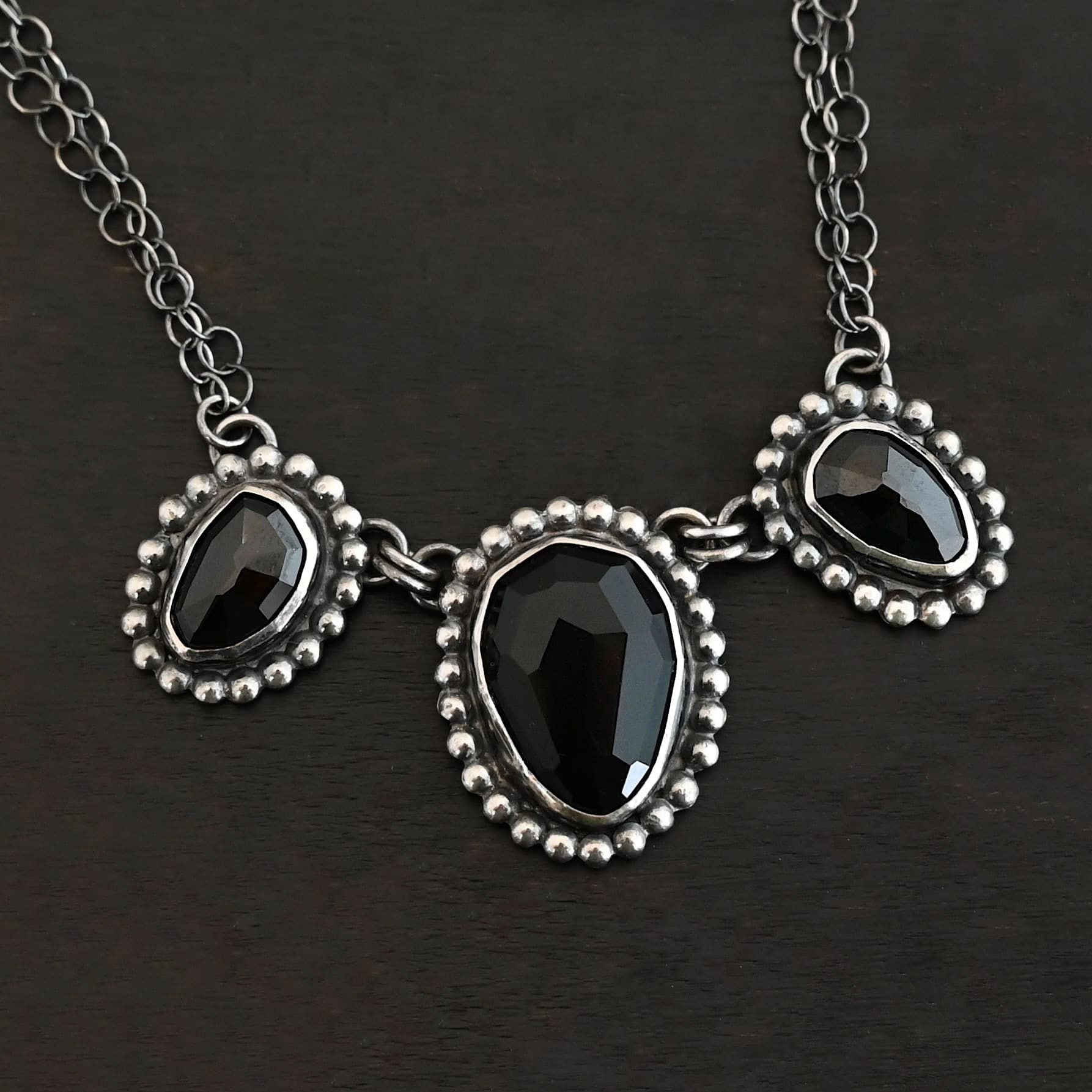 Close up of the "Escape To Tranquility" Necklace. Made out of sterling silver and three black onyx. The middle black onyx is the largest, with two smaller ones on the side. Each black onyx is set in silver and has silver balls going around it. Each black onyx is connected with two jump rings. Necklace is set against a dark background. Each side of the necklace is connected to two chains. Necklace is set against a dark background.
