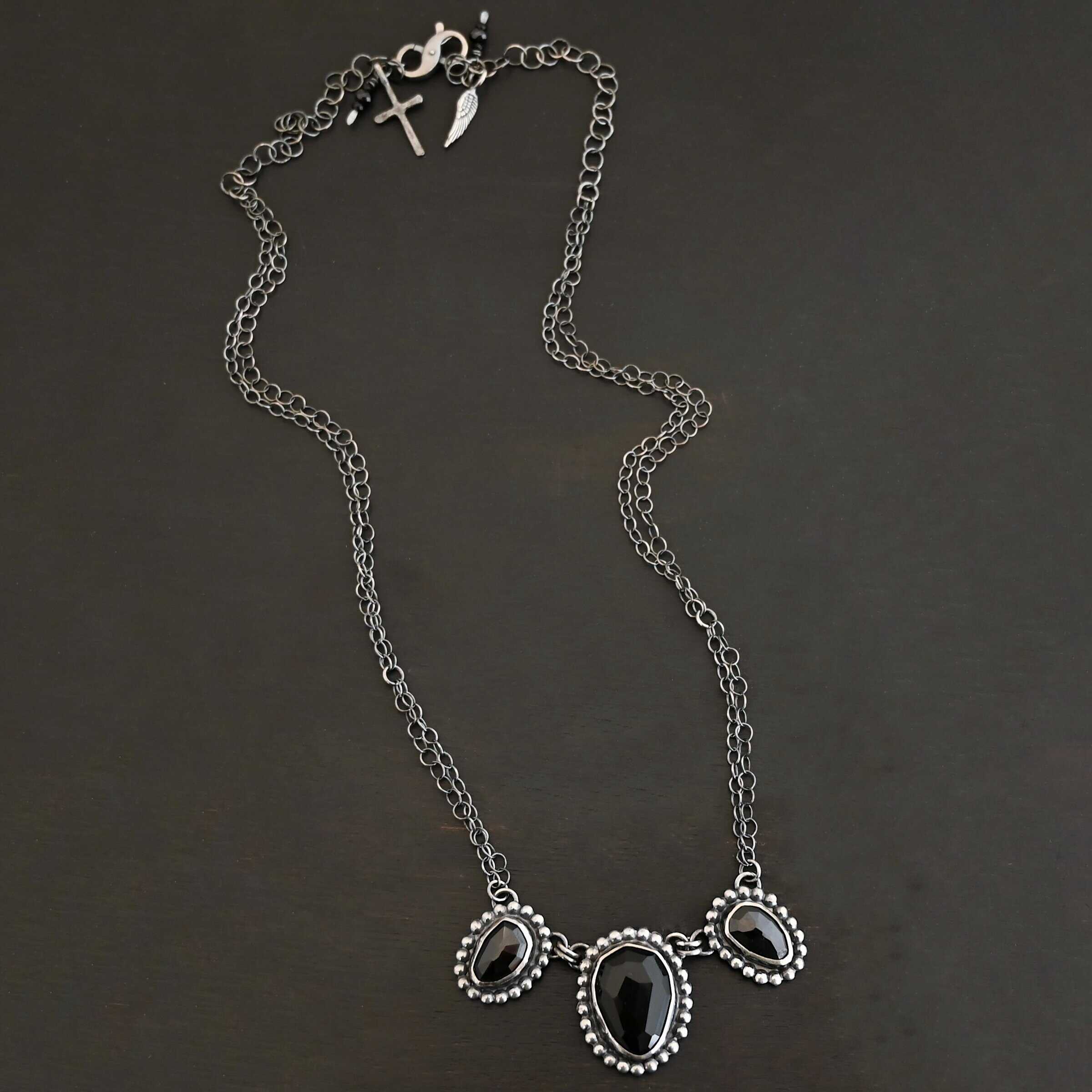 Close up of the "Escape To Tranquility" Necklace. Made out of sterling silver and three black onyx. The middle black onyx is the largest, with two smaller ones on the side. Each black onyx is set in silver and has silver balls going around it. Each black onyx is connected with two jump rings. Necklace is set against a dark background. Each side of the necklace is connected to two chains. Necklace is set against a dark background.