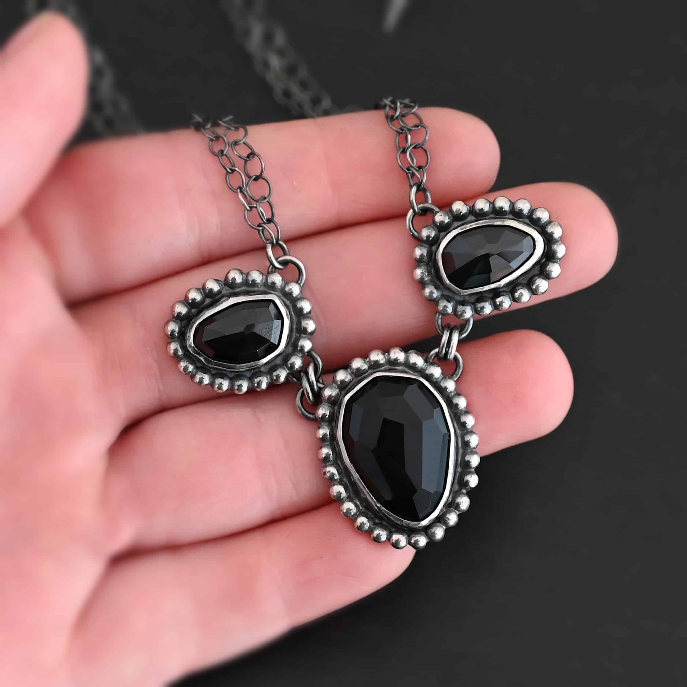 Close up of the "Escape To Tranquility" Necklace. Made out of sterling silver and three black onyx. The middle black onyx is the largest, with two smaller ones on the side. Each black onyx is set in silver and has silver balls going around it. Each black onyx is connected with two jump rings. Necklace is set against a dark background. Each side of the necklace is connected to two chains. Necklace is set against the palm of a hand to demonstrate size and length of the necklace.