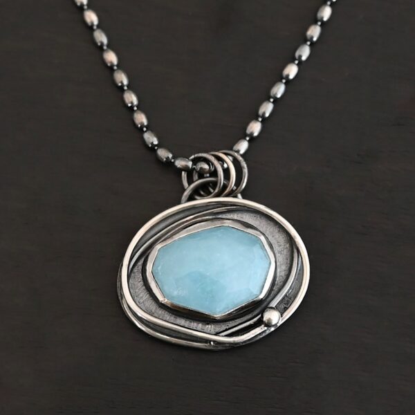 Close up of the "Marilee" Necklace. Made out of sterling silver and aquamarine. The aquamarine is in a slight oval shape and placed horizontally in the pendant. The silver overlaps as it circles the aquamarine, and there is one small silver dot in the bottom corner of the necklace. The necklace is on a "Bryn Beaded" Chain, which is made out of small oval beads. The necklace is laying against a dark background.