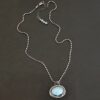 Close up of the "Marilee" Necklace. Made out of sterling silver & aquamarine. The aquamarine is an oval and set horizontally. There is a small silver dot in the bottom corner of the necklace, and the silver overlaps as it circles the aquamarine. The necklace is set against a dark background. The chain is a "Bryn Beaded" Chain, made out of small oval beads.