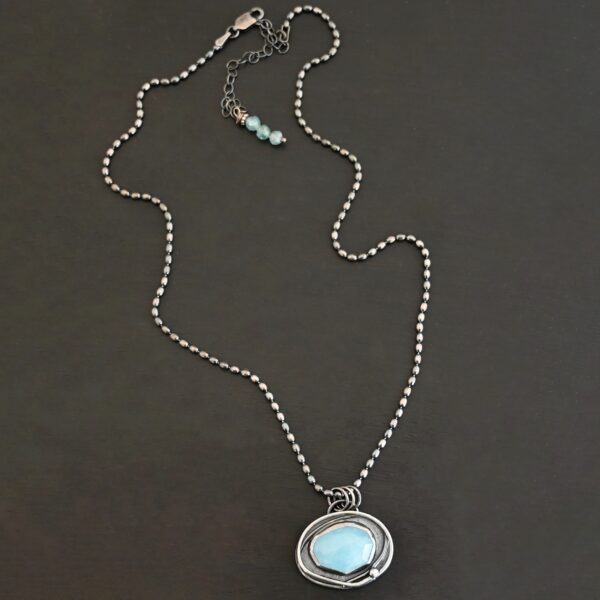 Close up of the "Marilee" Necklace. Made out of sterling silver & aquamarine. The aquamarine is an oval and set horizontally. There is a small silver dot in the bottom corner of the necklace, and the silver overlaps as it circles the aquamarine. The necklace is set against a dark background. The chain is a "Bryn Beaded" Chain, made out of small oval beads.