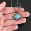Close up of the "Marilee" Necklace. Made out of sterling silver and aquamarine. The aquamarine is in a slight oval shape and placed horizontally in the pendant. The silver overlaps as it circles the aquamarine, and there is one small silver dot in the bottom corner of the necklace. The necklace is on a "Bryn Beaded" Chain, which is made out of small oval beads. The pendant is laying on a hand which is resting on a dark background.