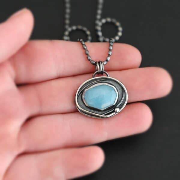 Close up of the "Marilee" Necklace. Made out of sterling silver and aquamarine. The aquamarine is in a slight oval shape and placed horizontally in the pendant. The silver overlaps as it circles the aquamarine, and there is one small silver dot in the bottom corner of the necklace. The necklace is on a "Bryn Beaded" Chain, which is made out of small oval beads. The pendant is laying on a hand which is resting on a dark background.