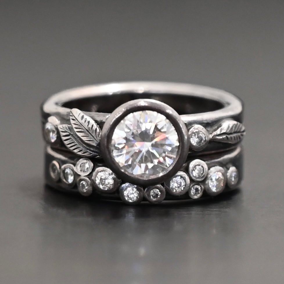 Stars In The Sky Ring "Stars In The Sky" Ring as shown up close. Made out of fifteen moissanites. The largest one is in the center on the top band. There is a small one on the right and another small one on the left side of the top band. There are three small engraved silver leaves on the top band, with two on the left and one on the right. The bottom band has twelve moissanites on it, all scattered. Ring is set against a dark background.