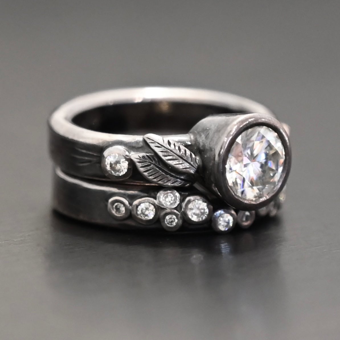 Stars In The Sky Ring "Stars In The Sky" Ring as shown up close. Made out of fifteen moissanites. The largest one is in the center on the top band. There is a small one on the right and another small one on the left side of the top band. There are three small engraved silver leaves on the top band, with two on the left and one on the right. The bottom band has twelve moissanites on it, all scattered. Ring is set against a dark background.