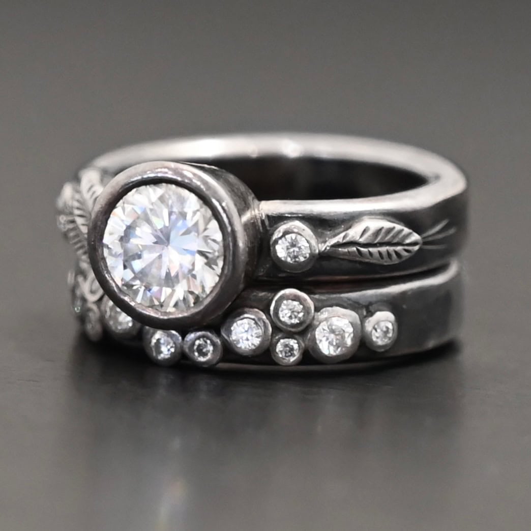 Stars In The Sky Ring "Stars In The Sky" Ring as shown up close. Made out of fifteen moissanites. The largest one is in the center on the top band. There is a small one on the right and another small one on the left side of the top band. There are three small engraved silver leaves on the top band, with two on the left and one on the right. The bottom band has twelve moissanites on it, all scattered. Ring is set against a dark background.