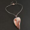"Rose In The Desert" Necklace. Made out of crazy lace agate and garnet. The crazy lace agate is the center stone. Pendant measures 2" by 3.5." It is a slight teardrop shape. The necklace has a curved bar on the left and right side that lead down to the pendant. There is a garnet in the center of where the bars meet before the pendant. Necklace is set against a dark background.