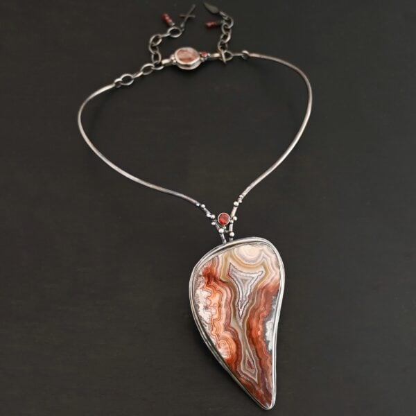 "Rose In The Desert" Necklace. Made out of crazy lace agate and garnet. The crazy lace agate is the center stone. Pendant measures 2" by 3.5." It is a slight teardrop shape. The necklace has a curved bar on the left and right side that lead down to the pendant. There is a garnet in the center of where the bars meet before the pendant. Necklace is set against a dark background.