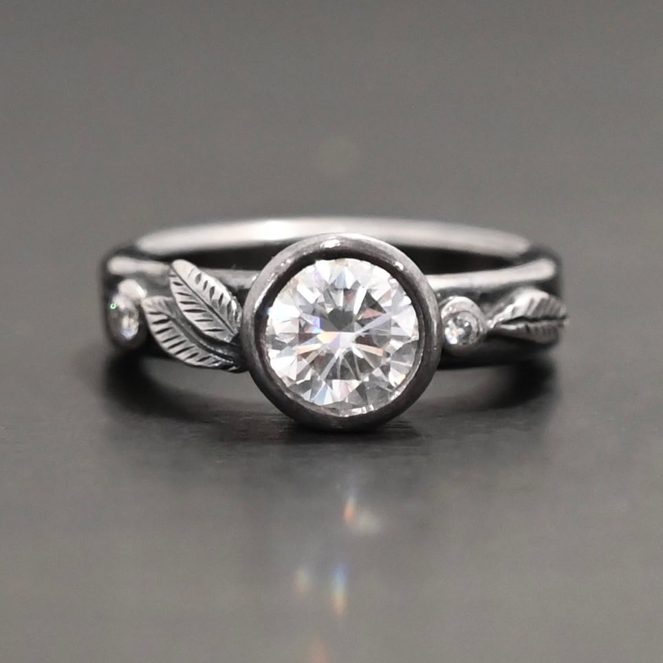 Stars In The Sky Ring "Stars In The Sky" Ring as shown up close. Made out of three moissanites. The largest one is in the center on the band. There is a small one on the right and another small one on the left side of the band. There are three small engraved silver leaves on the top band, with two on the left and one on the right.