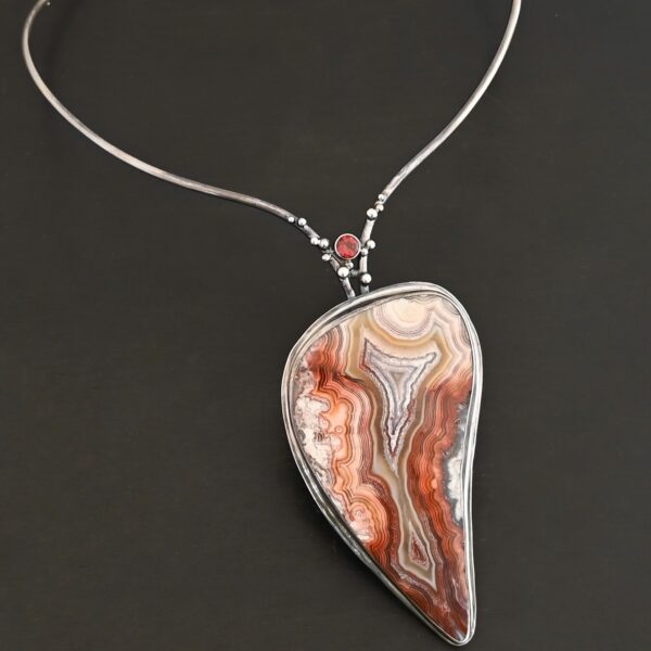 "Rose In The Desert" Necklace. Made out of crazy lace agate and garnet. The crazy lace agate is the center stone. Pendant measures 2" by 3.5." It is a slight teardrop shape. The necklace has a curved bar on the left and right side that lead down to the pendant. There is a garnet in the center of where the bars meet before the pendant. Necklace is set against a dark background.