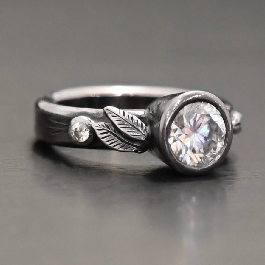 Stars In The Sky Ring "Stars In The Sky" Ring as shown up close. Made out of three moissanites. The largest one is in the center on the band. There is a small one on the right and another small one on the left side of the band. There are three small engraved silver leaves on the top band, with two on the left and one on the right.