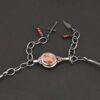 Close up of the "Rose In The Desert" clasp on the necklace. The clasp is made out of sterling silver and crazy lace agate and garnet. The crazy lace agate is circular and there is a small garnet on the end of the agate. There are three small silver dots on the other end of the clasp. There are three small garnet beads on both sides of the clasp, making for six beads. Clasp is set against a dark background. There is an angel wing pendant on the necklace as well.