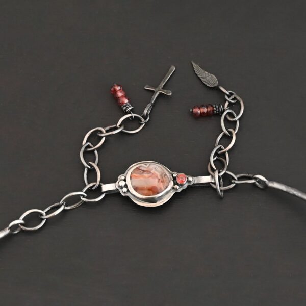 Close up of the "Rose In The Desert" clasp on the necklace. The clasp is made out of sterling silver and crazy lace agate and garnet. The crazy lace agate is circular and there is a small garnet on the end of the agate. There are three small silver dots on the other end of the clasp. There are three small garnet beads on both sides of the clasp, making for six beads. Clasp is set against a dark background. There is an angel wing pendant on the necklace as well.