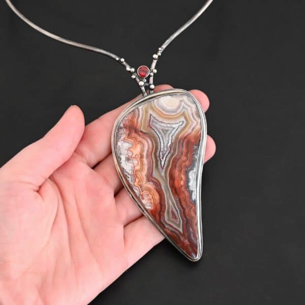 "Rose In The Desert" Necklace. Made out of crazy lace agate and garnet. The crazy lace agate is the center stone. Pendant measures 2" by 3.5." It is a slight teardrop shape. The necklace has a curved bar on the left and right side that lead down to the pendant. There is a garnet in the center of where the bars meet before the pendant. Necklace is set against the palm of a hand to demonstrate size and length of the necklace.