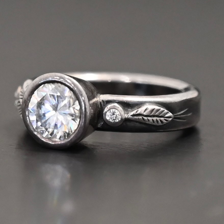 Stars In The Sky Ring "Stars In The Sky" Ring as shown up close. Made out of three moissanites. The largest one is in the center on the band. There is a small one on the right and another small one on the left side of the band. There are three small engraved silver leaves on the top band, with two on the left and one on the right.