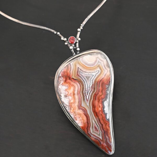 "Rose In The Desert" Necklace. Made out of crazy lace agate and garnet. The crazy lace agate is the center stone. Pendant measures 2" by 3.5." It is a slight teardrop shape. The necklace has a curved bar on the left and right side that lead down to the pendant. There is a garnet in the center of where the bars meet before the pendant. Necklace is set against a dark background.