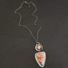 Close up of "The Greatest Journey" Necklace. This necklace is made out of Sterling Silver, Crazy Lace Agate, Garnet, & Citrine. The crazy lace agate is the biggest stone. It is a triangle shape. It is on the bottom of the necklace. It is a mix of brown, red, yellow, and orange swirls. It is set in silver, and around the stone setting is textured sterling silver. Above this stone is a large silver disk that contains a diamond shaped citrine that is rough textured. There is a small garnet that is a circle on the right side of the disk. There is a loop on the disk that attaches the pendant to the chain. Necklace is set against a dark background.