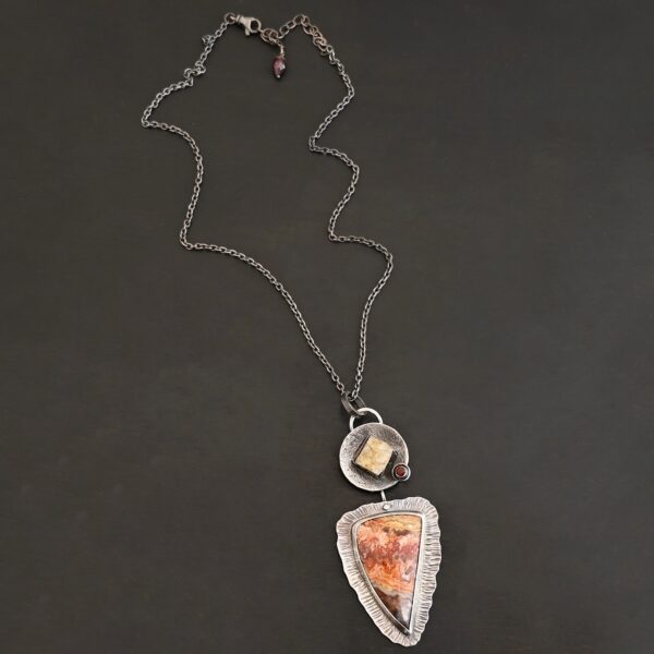 Close up of "The Greatest Journey" Necklace. This necklace is made out of Sterling Silver, Crazy Lace Agate, Garnet, & Citrine. The crazy lace agate is the biggest stone. It is a triangle shape. It is on the bottom of the necklace. It is a mix of brown, red, yellow, and orange swirls. It is set in silver, and around the stone setting is textured sterling silver. Above this stone is a large silver disk that contains a diamond shaped citrine that is rough textured. There is a small garnet that is a circle on the right side of the disk. There is a loop on the disk that attaches the pendant to the chain. Necklace is set against a dark background.