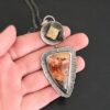 Close up of "The Greatest Journey" Necklace. This necklace is made out of Sterling Silver, Crazy Lace Agate, Garnet, & Citrine. The crazy lace agate is the biggest stone. It is a triangle shape. It is on the bottom of the necklace. It is a mix of brown, red, yellow, and orange swirls. It is set in silver, and around the stone setting is textured sterling silver. Above this stone is a large silver disk that contains a diamond shaped citrine that is rough textured. There is a small garnet that is a circle on the right side of the disk. There is a loop on the disk that attaches the pendant to the chain. Necklace is set against the palm of a hand to demonstrate size of the pendant.