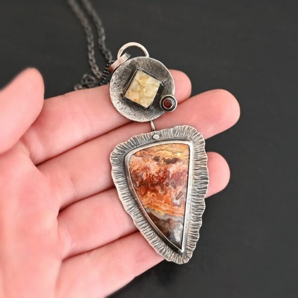 Close up of "The Greatest Journey" Necklace. This necklace is made out of Sterling Silver, Crazy Lace Agate, Garnet, & Citrine. The crazy lace agate is the biggest stone. It is a triangle shape. It is on the bottom of the necklace. It is a mix of brown, red, yellow, and orange swirls. It is set in silver, and around the stone setting is textured sterling silver. Above this stone is a large silver disk that contains a diamond shaped citrine that is rough textured. There is a small garnet that is a circle on the right side of the disk. There is a loop on the disk that attaches the pendant to the chain. Necklace is set against the palm of a hand to demonstrate size of the pendant.